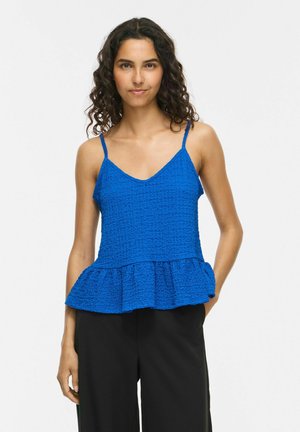 Femme aux cheveux bouclés portant un haut bleu texturé sans manches avec des fines bretelles et un pantalon noir, debout devant un fond clair uni.