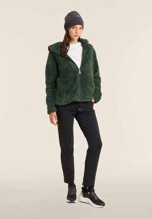 Giacca di pile verde con cappuccio, zip frontale e due tasche laterali. Abbinata a pantaloni neri e scarpe da ginnastica nere. Indossata con un berretto grigio.
