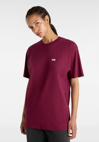 Jeune personne aux cheveux tressés portant un t-shirt Vans rouge foncé uni et un pantalon foncé sur fond blanc.