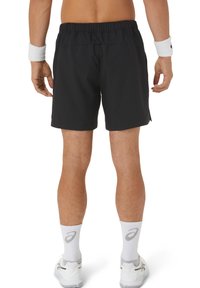 Schwarze Sportshorts mit elastischem Bund, knielang und mit seitlichen Schlitzen, getragen mit weißen Knöchelsocken und Turnschuhen.