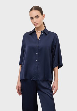 Donna che indossa una camicia di raso blu navy con bottoni e maniche a gomito, abbinata a pantaloni coordinati, posa davanti a uno sfondo grigio chiaro.