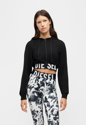 Mladá žena nosí čiernu krátku mikinu s textom "DIESEL" a čierno-biele tie-dye legíny na jednofarebnom pozadí.