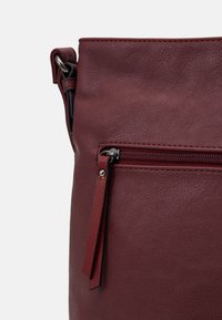 Bordeaux lædertaske med en tekstureret overflade, der har en lynlåslomme foran og en topklips. Udsmykket med sorte metaldele.