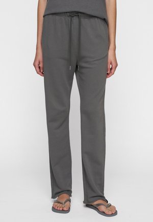 Persoon in losse grijze joggingbroek met rafelige zomen en grijze slippers, staand tegen een effen achtergrond.