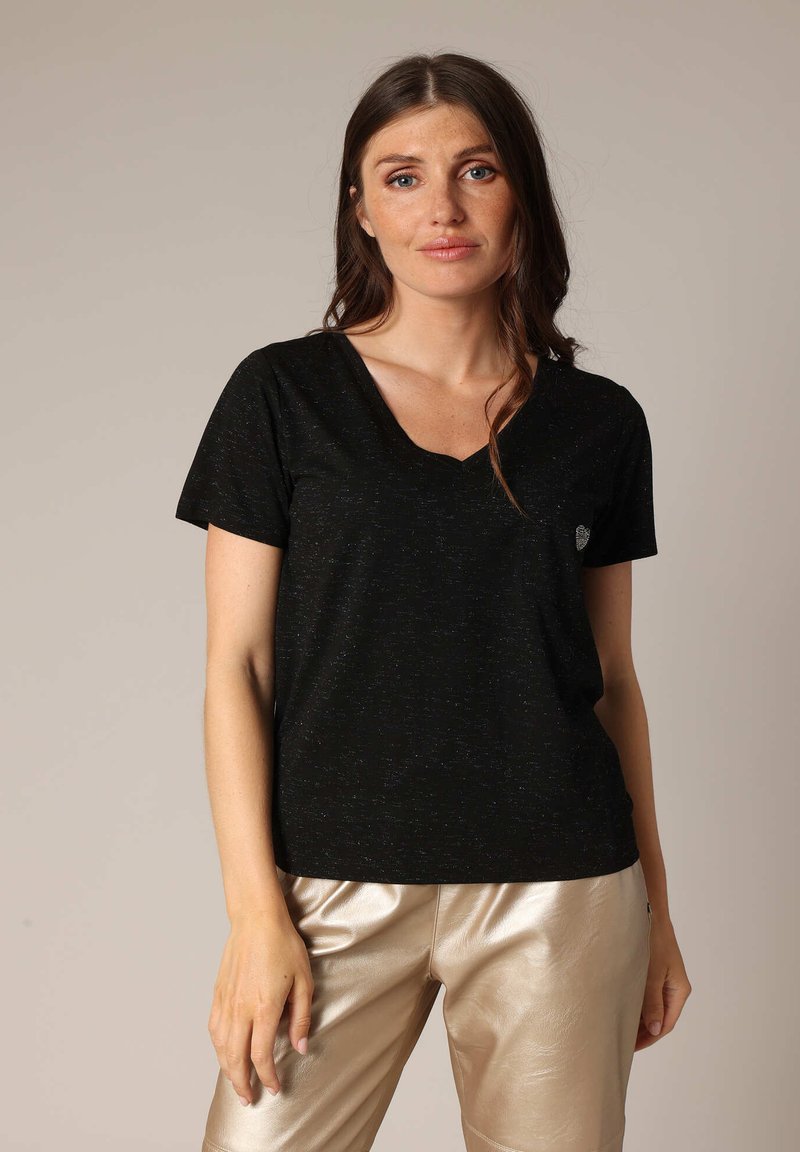 T-shirt noir à manches courtes en V avec un motif texturé, arborant un petit logo sur la poitrine et associé à un pantalon léger et brillant.