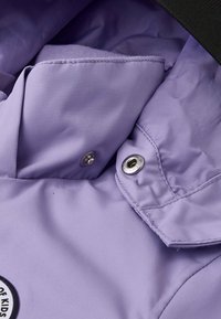 Giacca lilla con una texture morbida, chiusure a scatto e colletto nero a contrasto con una toppa rotonda.
