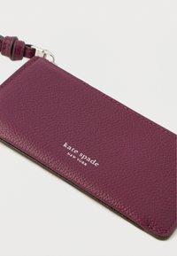 Fioletowy skórzany portfel na karty z teksturowaną powierzchnią, ozdobiony srebrnym logo i paskiem do przypięcia. Kompaktowy, prostokątny design.