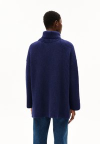 Marineblauer Rippstrick-Pullover mit Stehkragen, losem Schnitt und überschnittenen Schultern. Er hat einen mittellangen Schnitt und eine weiche Textur und wird mit blauen Jeans getragen.