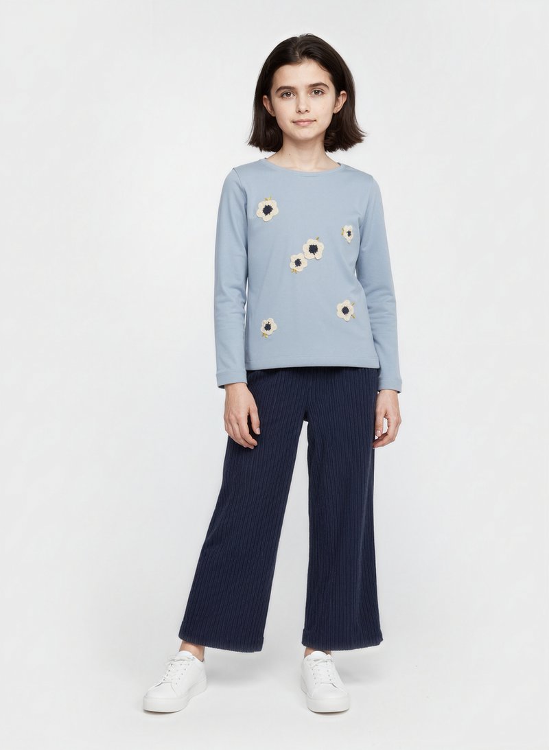 Hellblaues Langarm-Oberteil mit floralem Stickmuster und marineblauen, gerippten, weiten Hosen. Das Modell trägt weiße Sneakers und steht vor einem schlichten Hintergrund.