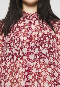 Blusa plissettata rossa con un colletto alto e arricciato. Presenta motivi floreali bianchi, tessuto semi-trasparente e bottoni sul collo.