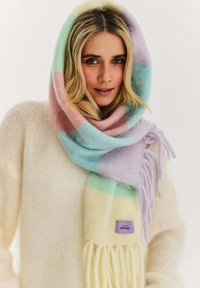 Kobieta o blond włosach, z pastelowym, paskowanym szalikiem owiniętym wokół głowy, ubrana w kremowy, puszysty sweter.