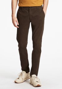 Sötétbarna, slim fit chino nadrág, lapos elejű és oldalsó zsebekkel, bézs sportcipőkkel párosítva, fehér fűzőkkel és mintás talppal.