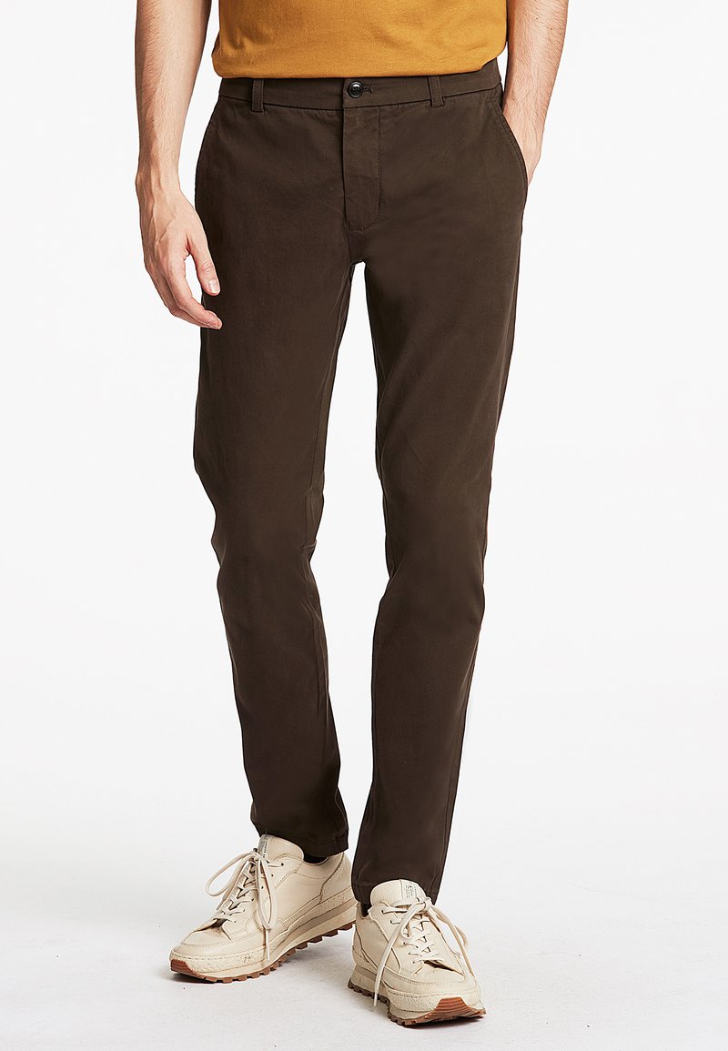 Sötétbarna, slim fit chino nadrág, lapos elejű és oldalsó zsebekkel, bézs sportcipőkkel párosítva, fehér fűzőkkel és mintás talppal.