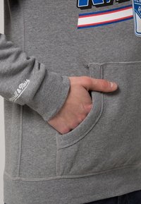 Sudadera gris con bolsillo tipo canguro, puños acanalados y el logo "Mitchell & Ness" bordado en la manga. Tela suave con una textura lisa.