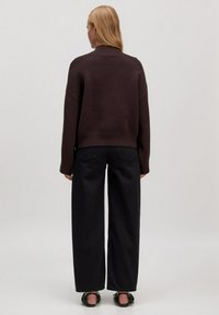 Braun gestrickter Pullover mit hohem Kragen, kombiniert mit lockeren schwarzen Hosen und schwarzen Absatzsandalen. Glatte Textur, minimales Design.
