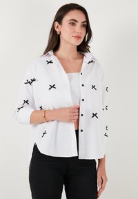 White button-up shirt with black embroidered X patterns, a collared neck, cuffed sleeves, and a relaxed fit. Smooth, lightweight fabric.