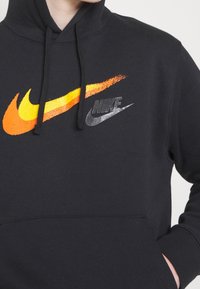 Svart hoodie med två Nike-logotyper: en gradient orange och gul swoosh samt en metallisk silverfärgad swoosh. Dragskohuva och framficka.