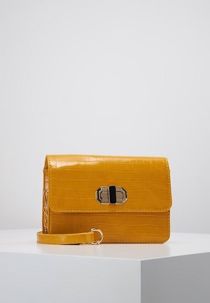 Petit sac bandoulière jaune moutarde avec texture crocodile, fermoir tourniquet doré et bandoulière poignet amovible sur surface blanche.