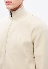 Bézs színű cipzáros dzseki, magas gallérral, sima anyagból készült, a mellrészen tónusos "BOSS" logóval, és testhezálló kialakítással.