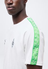 Άνδρας που φοράει λευκό μπλουζάκι με πράσινη ριγέ μοτίβο στον ώμο και μαύρο διαμαντένιο λογότυπο στο στήθος, πάνω σε απλό φόντο.