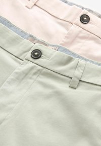 Pantalones de tela verde claro y rosa pálido con botones negros, diseño de frente plano y textura sutil. Con un forro de patrón a cuadros.