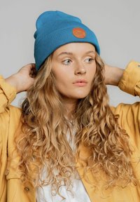 Niebieska, prążkowana czapka beanie z okrągłą, brązową skórzaną łatą. Model nosi żółtą kurtkę na białej koszuli, podkreślony kręconymi włosami.