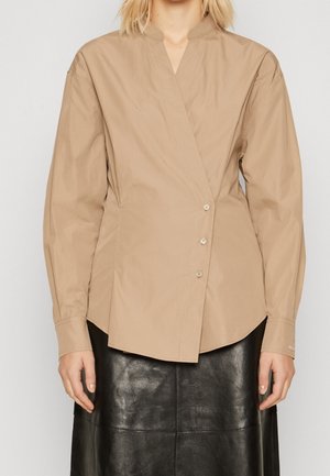 Kvinde iført en beige asymmetrisk knappet bluse med lange ærmer og et sort lædernederdel.