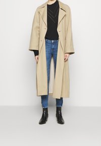 Beige lang frakke med brede revers, sort sweater med struktureret mønster, blå skinny jeans og sorte ankelstøvler med blank finish.