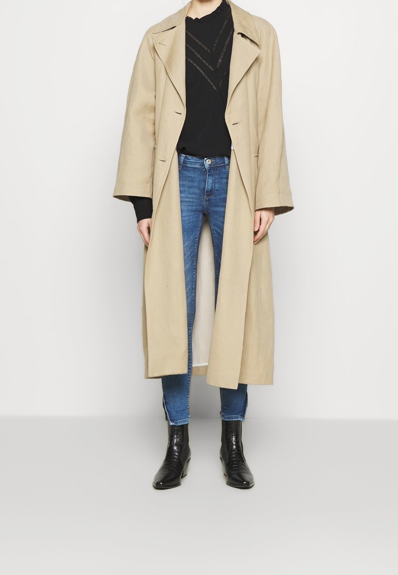 Beige lang frakke med brede revers, sort sweater med struktureret mønster, blå skinny jeans og sorte ankelstøvler med blank finish.