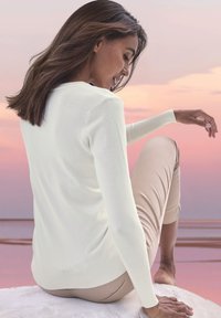 Weißer Pullover mit langen Ärmeln und geripptem Saum, kombiniert mit hellbeigen, kurz geschnittenen Hosen. Die Textur wirkt glatt und weich.