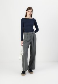 Top azul marinho de manga longa com decote quadrado, combinado com calças cinzentas de cintura alta e pregas. A modelo leva uma mala branca e usa sapatilhas pretas.