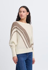 Donna con lunghi capelli scuri che indossa un maglione color crema con strisce diagonali marroni e jeans blu, che guarda a sinistra su uno sfondo semplice.