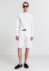 Camicetta bianca a maniche lunghe con colletto alla coreana, abbinata a pantaloni corti bianchi e mocassini scuri. Gli accessori includono occhiali da sole neri. Texture del tessuto liscia.