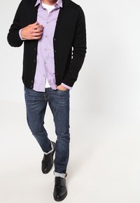 Cardigan nero sopra una camicia button-up viola chiaro, jeans scuri e scarpe nere. L'outfit presenta un design casual con dettagli dei bottoni.