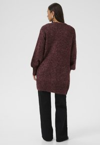 Oversized burgundy knit sweater with a textured finish, ribbed hem, and long sleeves. Worn with black trousers, view from the back.