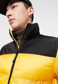 Gelbe und schwarze Daunenjacke mit hohem Kragen. Verfügt über einen Reißverschluss und eine gerippte Textur am Kragen. Leichtes, gepolstertes Design.