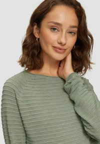 Haut à manches longues vert rayé avec un décolleté rond et un tissu texturé. Le modèle touche son cou, mettant en avant le design de manches adaptable.