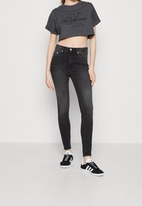 Jeans ajustados de cintura alta en negro combinados con una camiseta corta gris que muestra un logo negro. La modelo lleva zapatillas negras y blancas.