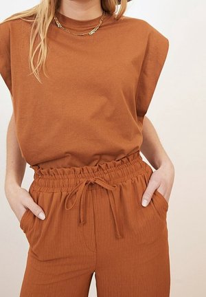 Vrouw draagt een roestkleurige mouwloze top en een broek met hoge taille en koordje, handen in de zakken en een gouden ketting.