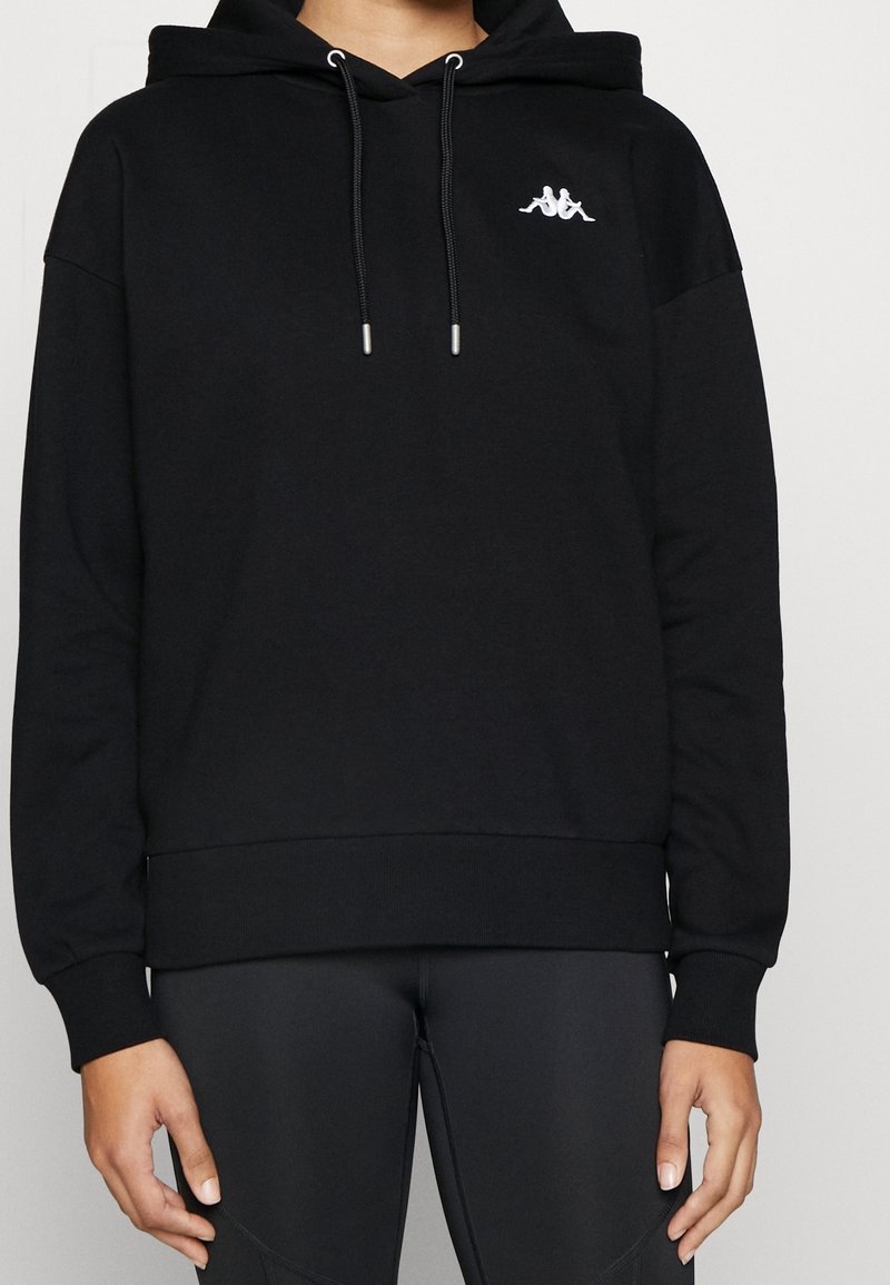 Persona che indossa un hoodie nero con un logo bianco sul petto e leggings neri abbinati, in piedi con le mani rilassate ai lati.