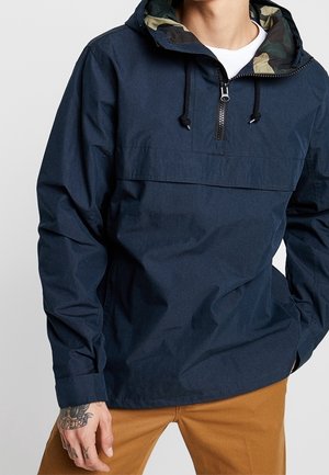 Personne portant une veste à enfiler bleu marine avec une demi-fermeture éclair et une capuche doublée camouflage, associée à un pantalon marron, main posée près de la hanche.