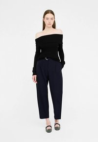 Haut noir à manches longues et épaules dénudées, associé à un pantalon taille haute bleu marine. Le mannequin est debout, les mains dans les poches, portant des ballerines noires.