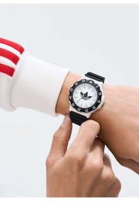 Must mustriba kell musta kummirihmaga, valge põhja, musta raamiga ja mustade aktsentidega; sisaldab Adidas'e logo ja suuri numbreid, et oleks lihtne lugeda.