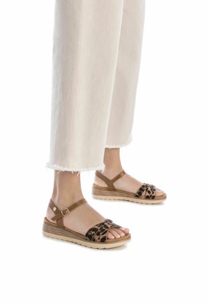 Sandalias marrones con correas de estampado de leopardo, una suela acolchada y una correa en el tobillo ajustable, combinadas con pantalones blancos de tiro deshilachado.