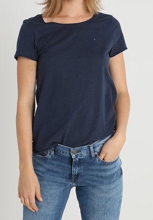 Kvinde iført en ensfarvet mørkeblå t-shirt med korte ærmer, let stoppet ind i midtalje blå jeans, med den ene hånd i lommen, foran en ensartet baggrund.