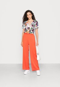 Top de manga curta floral com padrão tropical multicolorido, combinado com calças largas, de cintura alta, laranja e ténis brancos.