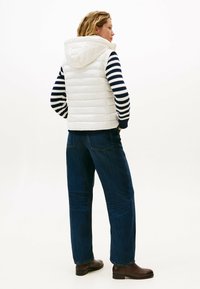 Gilet imbottito bianco trapuntato con cappuccio, indossato sopra un maglione a righe blu navy e bianco, abbinato a jeans blu scuro e stivaletti marroni.