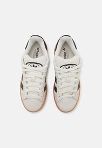Zapatillas de ante blancas con rayas negras, con cordones beige y suela de goma. Logo de Adidas visible en la lengüeta.