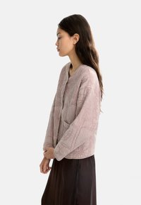 Kobieta w profilu bocznym, ubrana w jasnoróżową, strukturalną kurtkę i ciemnobrązową spódnicę, stojąca na tle jednolitego, białego tła.