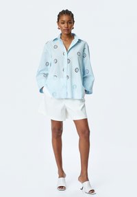 Hellblaues Button-Up-Hemd mit floralen Ausschnitten, langen Ärmeln, kombiniert mit weißen Shorts und weißen Schlupfschuhen. Glatte Textur.